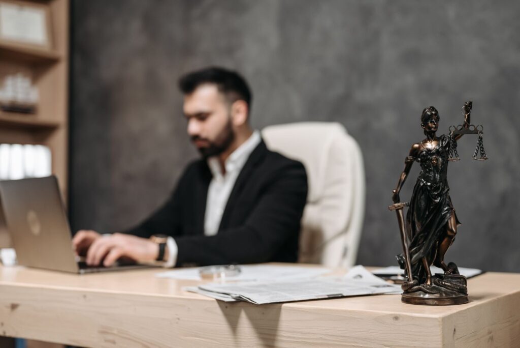 A businessman working on a laptop in an office with a Lady Justice statue in focus on the desk.
